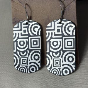 May include: A pair of black and white geometric pattern earrings. The earrings are rectangular with rounded tops and feature a repeating design of circles and squares. The earrings hang from silver-colored hooks.