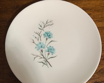 May include: A white ceramic plate with a floral design. The plate features a cluster of blue flowers with green stems and leaves. The flowers are in the center of the plate. The plate is on a dark brown wooden surface.