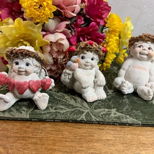 May include: Three cherub figurines with wings and halos. One holds a banner of red hearts, another has a bird on its shoulder, and the third is seated. They are on a green patterned cloth, with a colorful flower arrangement in the background.