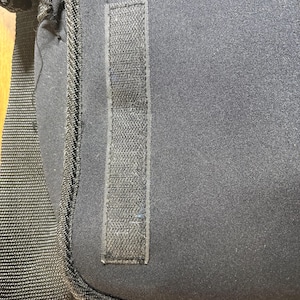 May include: Close-up of a black shoulder bag. The bag features a textured, rectangular Velcro strip on the front. The bag has a black strap and a black zipper. The bag is made of a soft, durable material.
