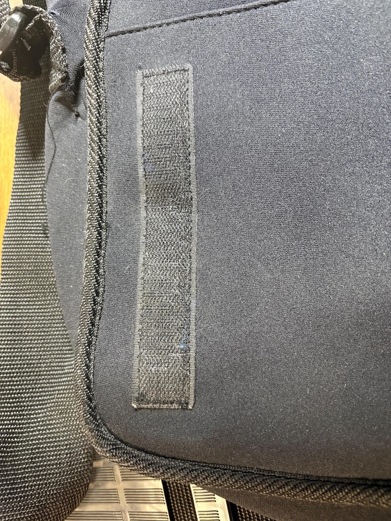 May include: Close-up of a black shoulder bag. The bag features a textured, rectangular Velcro strip on the front. The bag has a black strap and a black zipper. The bag is made of a soft, durable material.