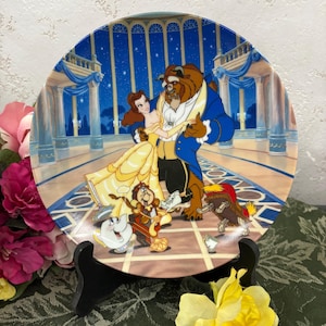 May include: A ceramic plate depicting a scene from the Disney film Beauty and the Beast. Belle and the Beast are dancing in a grand ballroom, surrounded by other characters from the film, including Lumiere, Cogsworth, and Mrs. Potts. The plate is decorated with a blue and gold design.