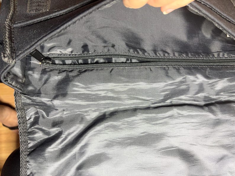 May include: Black bag interior with a silver-colored, shiny lining. A black zipper runs across the top, with a black zipper pull. The bag's exterior is black, and the image is taken from an overhead perspective.