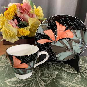 May include: A black and white teacup and saucer set with a floral design. The cup and saucer feature a black background with a large, orange flower and green leaves.