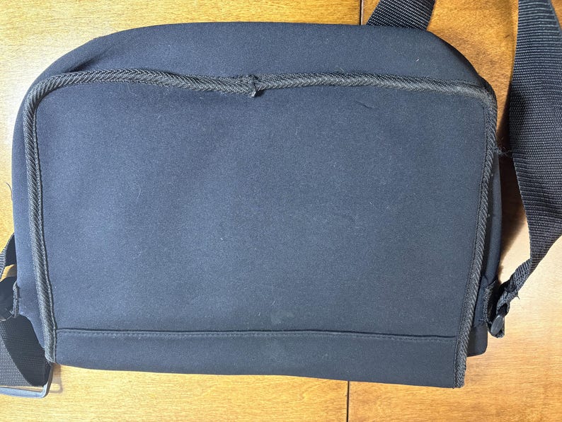 May include: A black messenger bag with a shoulder strap. The bag is made of a dark, possibly neoprene-like material. The bag has a rectangular shape with rounded corners and a zipper closure. The shoulder strap is black.