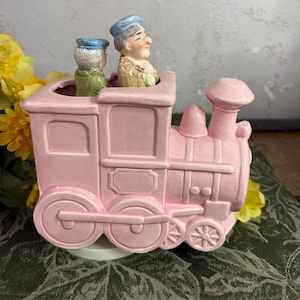 May include: A pink ceramic train engine with two figures of people wearing blue hats riding on top. The train has black wheels and a white base.