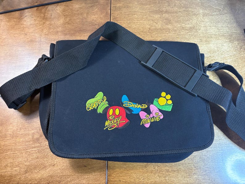 May include: Black messenger bag with a black adjustable shoulder strap. The bag features colorful embroidered patches of Disney characters including Mickey, Minnie, Donald, and Goofy. The bag is made of a durable, water-resistant material.