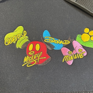 May include: Embroidered patches featuring Disney characters Goofy, Mickey, Donald, and Minnie. Each patch has a unique design and color scheme, with the character's name in yellow script.