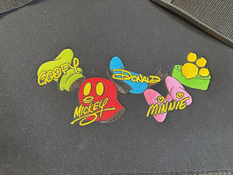 May include: Embroidered patches featuring Disney characters Goofy, Mickey, Donald, and Minnie. Each patch has a unique design and color scheme, with the character's name in yellow script.