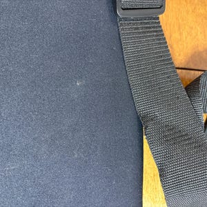 May include: Black fabric with a black adjustable strap and a black plastic buckle. The strap is made of a woven material. The background is a light brown wooden surface.