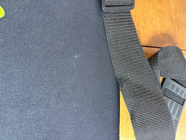 May include: Black fabric with a black adjustable strap and a black plastic buckle. The strap is made of a woven material. The background is a light brown wooden surface.