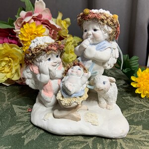 Puede incluir: Una figurita de cerámica blanca que representa una escena de la natividad con dos ángeles, un bebé Jesús en un pesebre y un cordero blanco. Los ángeles llevan coronas de flores y tienen alas. El bebé Jesús está envuelto en una manta azul y está acostado en un pesebre hecho de paja. El cordero está de pie junto al pesebre.