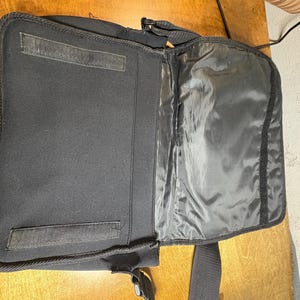 May include: Open black laptop bag with a shoulder strap. The bag is made of a soft, padded material and features a large main compartment and a smaller pocket. The bag has a velcro closure and a zipper.