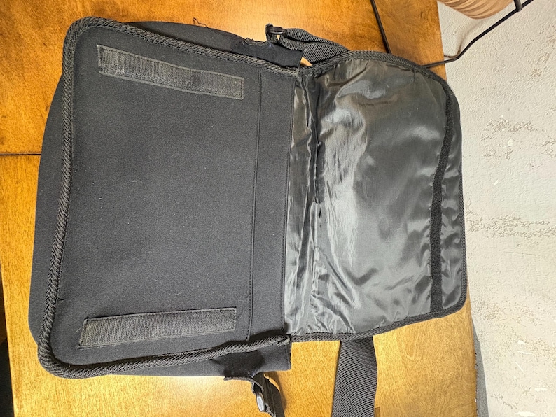 May include: Open black laptop bag with a shoulder strap. The bag is made of a soft, padded material and features a large main compartment and a smaller pocket. The bag has a velcro closure and a zipper.