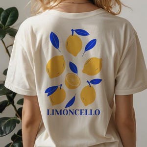 May include: A cream-coloured t-shirt with a lemon design and the word "LIMONCELLO" in blue. The graphic features yellow lemons and blue leaves. The shirt has short sleeves and a relaxed fit, perfect for casual wear. The design is on the back.
