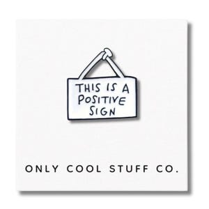 May include: A white enamel pin with a black outline, shaped like a sign. The pin features the text "THIS IS A POSITIVE SIGN" in black. Below the pin, the text reads "ONLY COOL STUFF CO.".