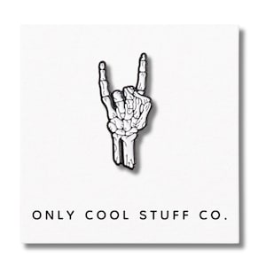 Puede incluir: Un pin de esmalte en blanco y negro que representa una mano de esqueleto haciendo el gesto de rock and roll. El pin está sobre un fondo blanco con el texto "ONLY COOL STUFF CO." en la parte inferior.