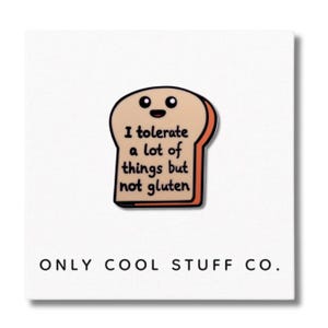 May include: A white enamel pin shaped like a slice of bread with a smiling face and the text "I tolerate a lot of things but not gluten". The pin is on a white background with the text "ONLY COOL STUFF CO."