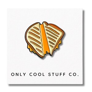 Puede incluir: Un pin de esmalte amarillo y marrón de un sándwich de queso a la parrilla con un bocado sacado. El pin dice "ONLY COOL STUFF CO."