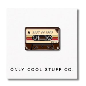 May include: A brown and beige cassette tape pin with the text "BEST OF 1980" and "SU 90" on it. The pin is on a white background with the text "ONLY COOL STUFF CO."