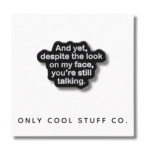May include: A black enamel pin featuring the phrase "And yet, despite the look on my face, you're still talking." The pin is in a speech bubble shape, with the words "ONLY COOL STUFF CO." at the bottom.