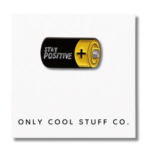 Puede incluir: Un pin de esmalte con forma de batería negra y amarilla con las palabras "STAY POSITIVE" y un signo más. El pin está en una tarjeta blanca con el texto "ONLY COOL STUFF CO."
