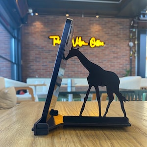 May include: A black metal phone stand with a giraffe silhouette design. The stand is holding a black smartphone.