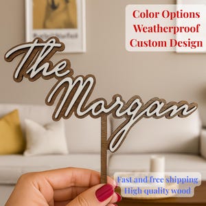 Op de afbeelding: Een houten taarttopper met de woorden "The Morgan" in een gelaagd ontwerp. De bovenste laag is wit en de onderste laag is donkerbruin. De tekst "Color Options, Weatherproof, Custom Design" is zichtbaar.