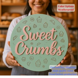 May include: A circular sign in mint green, featuring the pink text "Sweet Crumbs". The sign is adorned with line drawings of cupcakes and pastries. The sign is held by a person. The sign is weatherproof.