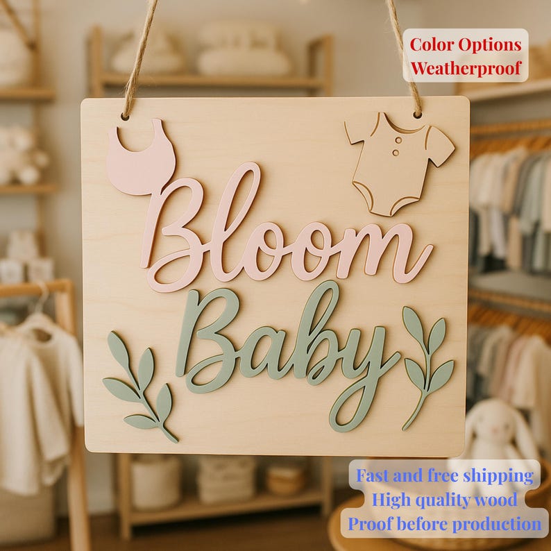 May include: A wooden sign featuring the words "Bloom Baby" in pink and green script, with a bib, babygrow, and leaf details. The sign is made of light-coloured wood and is designed to be hung. The text "Color Options Weatherproof" is also visible.
