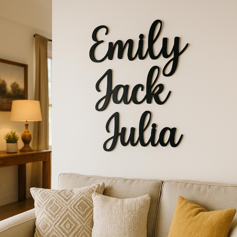 Op de afbeelding: Zwarte houten wanddecoratie met de namen "Emily", "Jack" en "Julia" in een sierlijk lettertype. De namen zijn bevestigd aan een witte muur boven een beige bank met decoratieve kussens. Een lamp en kunstwerk zijn zichtbaar op de achtergrond.