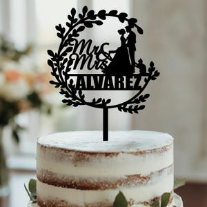 May include: Black silhouette of a couple with a wreath of leaves around them. The wreath has the words "Mr & Mrs ALVAREZ" written on it. The silhouette is on a cake topper.