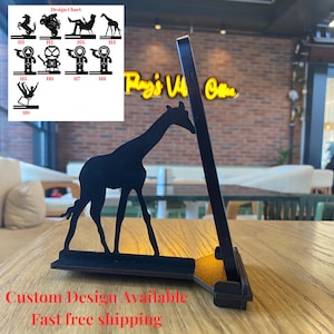 May include: A black metal phone stand with a giraffe silhouette design. The stand is on a wooden table. The text "Custom Design Available" and "Fast free shipping" is visible at the bottom of the image.