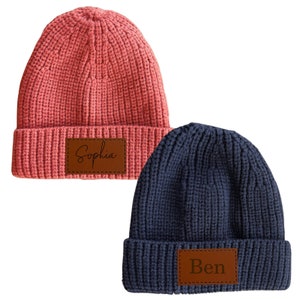 May include: Two knitted beanies with personalized leather patches. The beanie on the left is pink and says "Sophia". The beanie on the right is blue and says "Ben".
