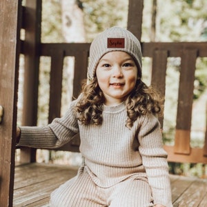 May include: A young child wearing a beige knitted sweater and matching pants set. The child is wearing a beige beanie with a brown leather patch that says "Sophia".