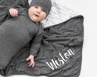 May include: A dark gray long-sleeve baby gown and matching beanie cap set. The set includes a dark gray blanket with a white textured dimpled underside. The blanket features the name Weston printed in white script lettering on the dark gray side.