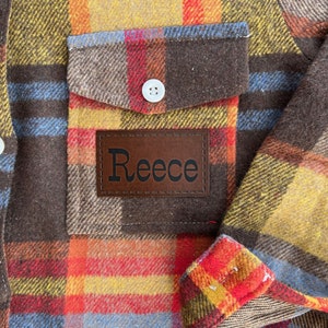 May include: A brown, yellow, red, and blue plaid flannel shirt with a brown leather patch that says "Reece" on the front left pocket.