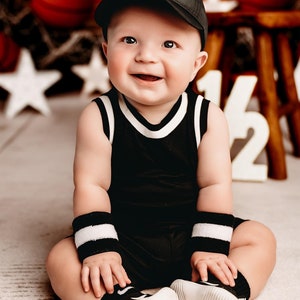 May include: A baby boy wearing a black and white basketball jersey, a black baseball cap with a white logo, and white and black wristbands. The baby is sitting on a white surface with a basketball theme background.