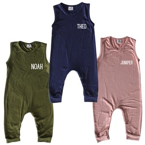 May include: Three sleeveless baby rompers in olive green, navy blue, and dusty rose. Each romper has a name printed on the front: "NOAH", "THEO", and "JUNIPER". The rompers have a round neckline and snap closures at the crotch.