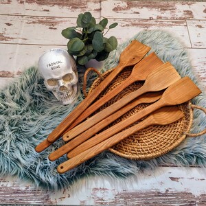May include: A set of six wooden kitchen spatulas with natural wood grain. The spatulas are arranged in a fan shape on a woven wicker tray. The spatulas are all the same size and shape, and they have a long, thin handle.