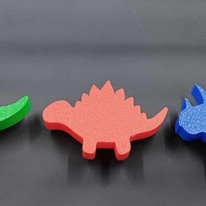 May include: Three foam dinosaur shapes, a green Tyrannosaurus Rex, a red Stegosaurus, and a blue Triceratops.