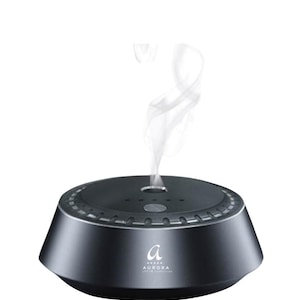 Peut inclure: Un diffuseur d'huiles essentielles circulaire noir mat avec une base conique et un dessus plat. Un mince filet de brume blanche s'échappe de l'évent central. L'avant présente un logo blanc avec la lettre a et le texte AURORA en dessous.