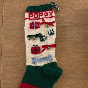 May include: A hand-knitted Christmas stocking with the name "POPPY" at the top. The stocking features red, green, and white sections, with gift and paw print designs. Small bells and bows adorn the front, and a blue bone is also present.