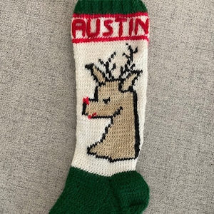 May include: A knitted Christmas stocking with a green, white and red colour scheme. The stocking features a reindeer design and the name "Austin" embroidered on the top.