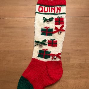 May include: A handmade Christmas stocking with the name "QUINN" knitted in red letters. The stocking is primarily white with red and green accents, featuring gift box designs and small bells. The toe and heel are green.