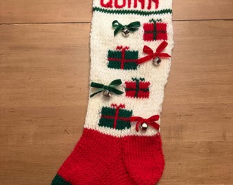 May include: A knitted Christmas stocking, predominantly white with red and green detailing. The name "QUINN" is displayed in red. Decorated with gift box motifs, small bells, and a green toe and heel. Festive and handmade.