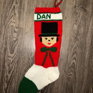 May include: Red and green knitted Christmas stocking with a white cuff and a cartoon face with a black top hat. The stocking is personalized with the name "DAN".