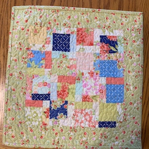 May include: A green quilted square with a patchwork design featuring floral and geometric patterns in shades of pink, blue, yellow, and green.