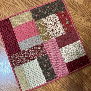 May include: A handmade patchwork quilt with a variety of floral and dotted patterns in shades of red, pink, beige, and brown. The quilt is square-shaped and has a pink border.