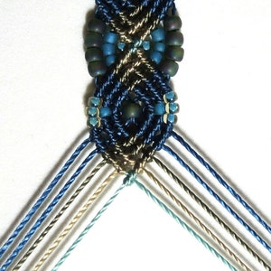 May include: A close-up of a blue, brown, and green macrame bracelet with beads. The bracelet is made with a variety of different colored cords and beads.